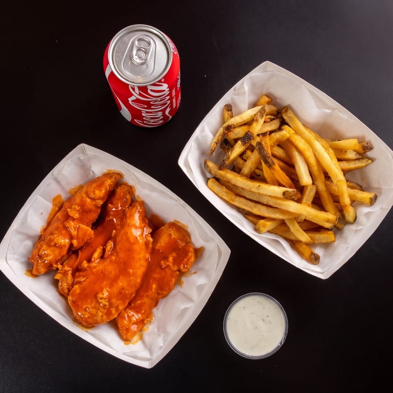 Top-down view of buffalo chicken tenders, fries, a Coca-Cola can, and ranch dressing on a black surface.