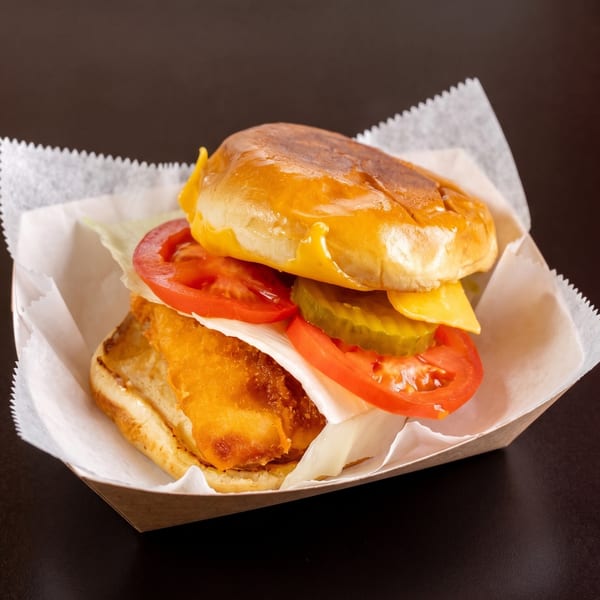 Crispy fish sandwich with lettuce, tomato, pickles, and cheese on a brioche bun in a paper tray.